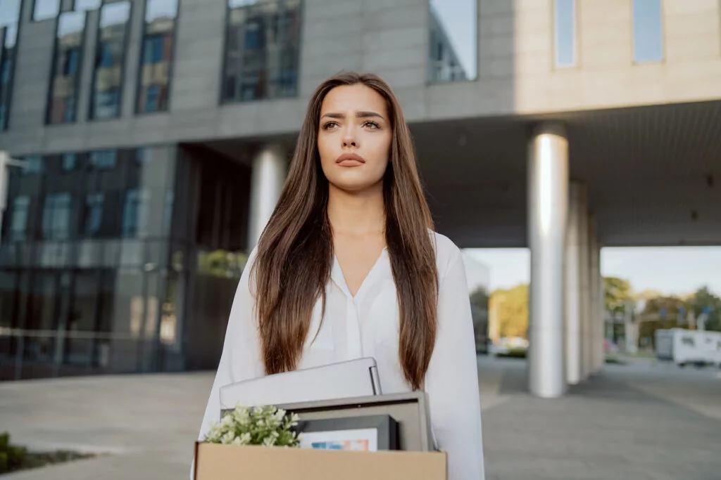 Woman Stands In Front Of Glass Modern Building Of Corporation Office Where She Worked Dismissal From Position Girl With Sad Uncertain Face Looks Ahead Holding Box With Packed Things Unemployment for article on signs you should quit your job
