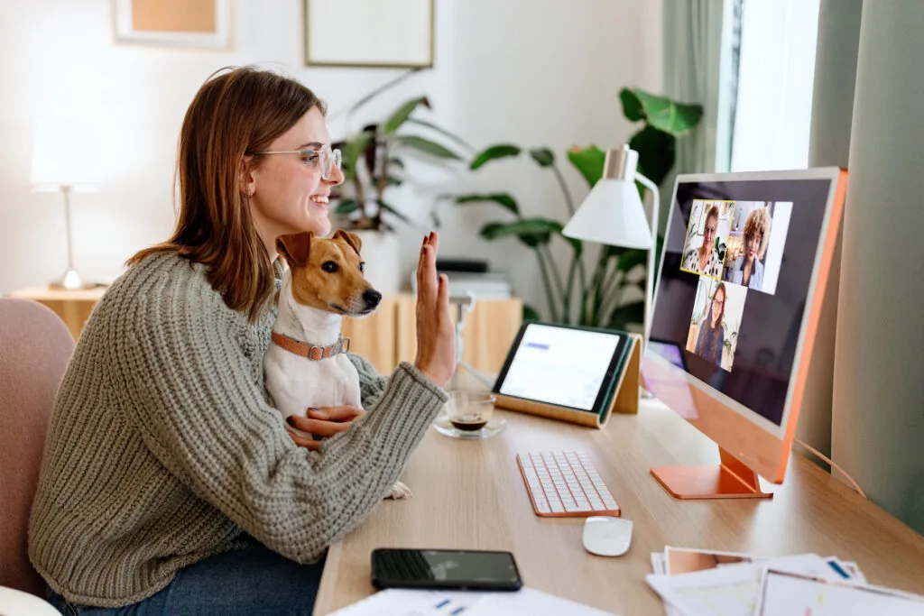 Woman working hybrid job sitting at desk with her dog in front of computer for remote virtual work meeting