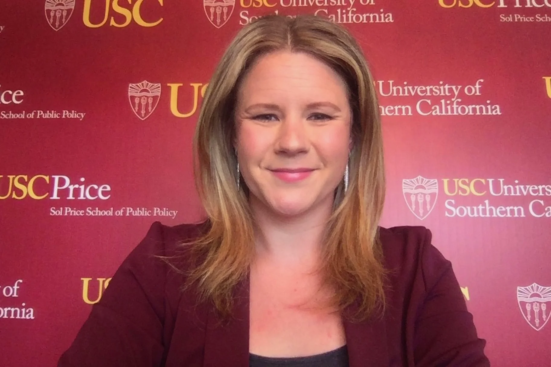 Read about: USC Price online alumna Kate Kelly smiles in red blazer in front of USC step and repeat banner.