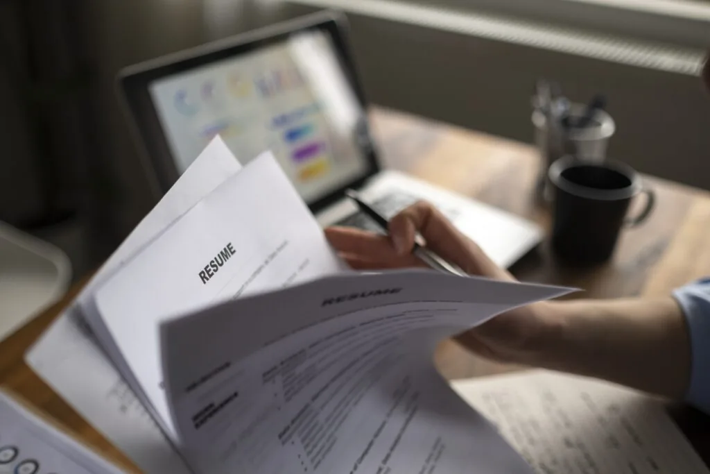 Recruiter's hands leaf through pile of resumes in front of a laptop. Learn how to avoid key resume mistakes in this article.