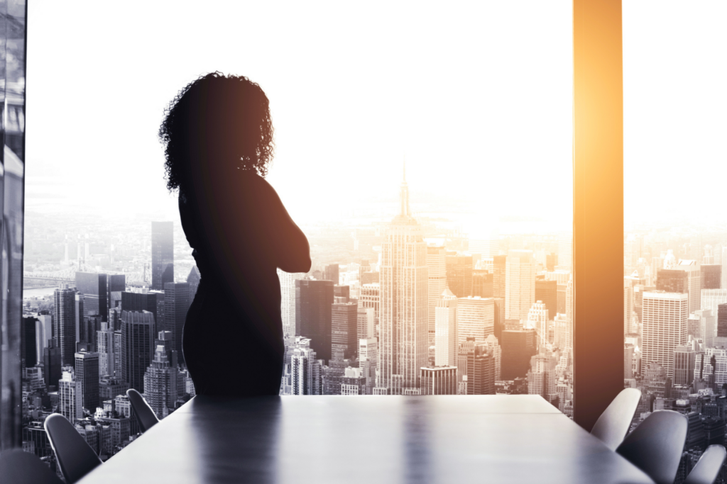 A woman looks out the window of a boardroom at a city skyline at sunset.