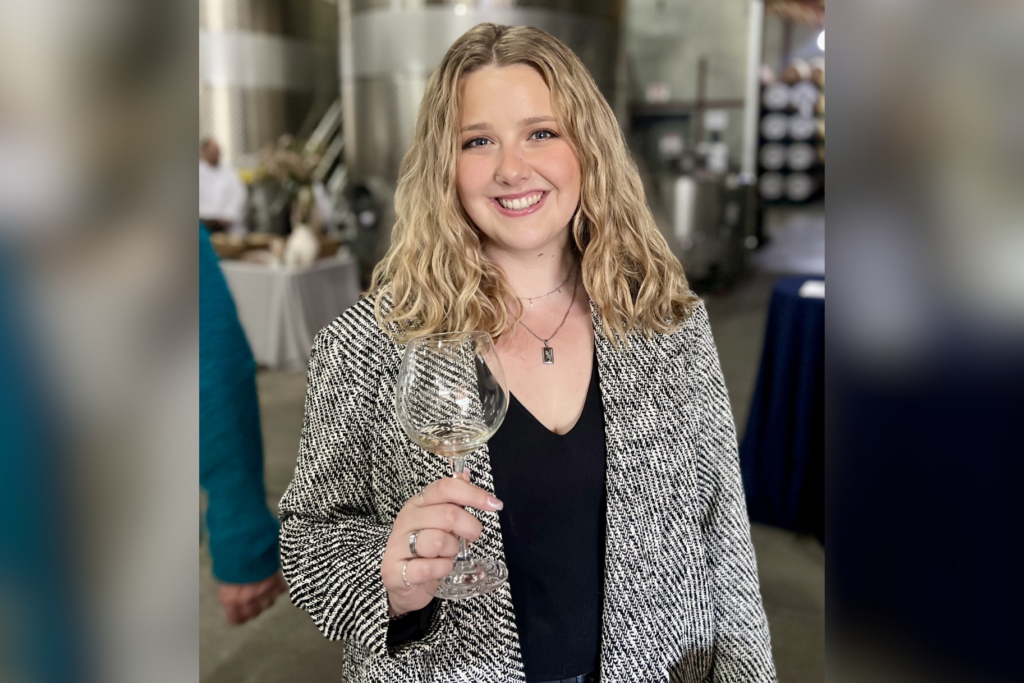 Emily Atkinson stands in a winery with cement walls holding a glass of white wine.