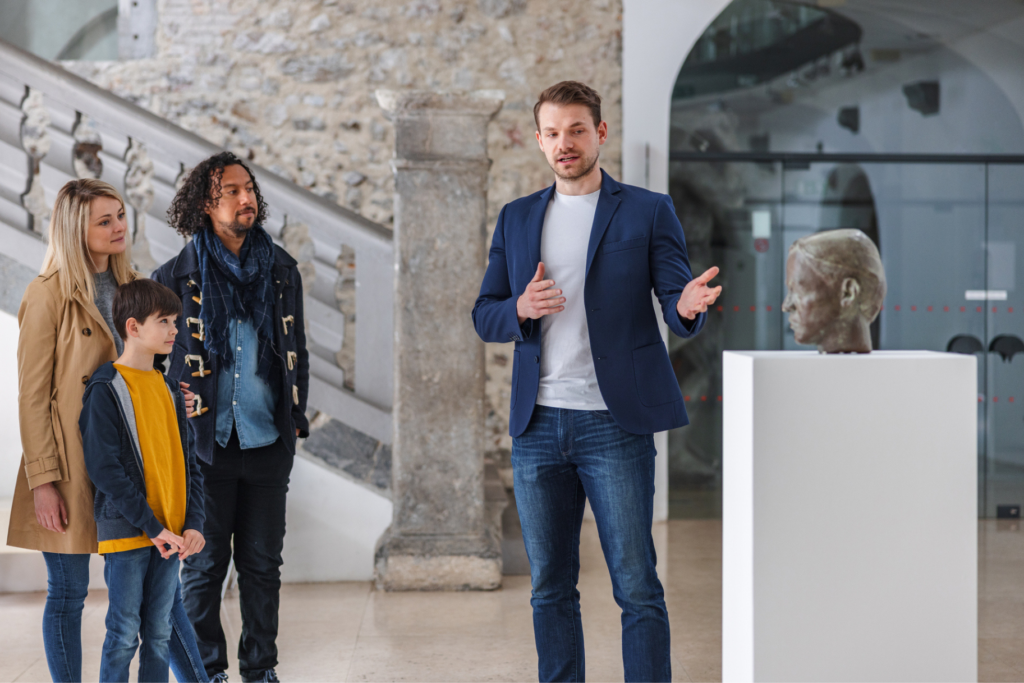 Man gestures to sculpture while giving tour to family in museum. In this article, learn how to be a museum curator and explore other jobs for creative people.