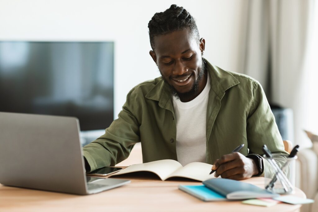 Smiling African American Man Sitting At Desk, Working On Laptop And Taking Notes In Notebook, Black Male Studying Online