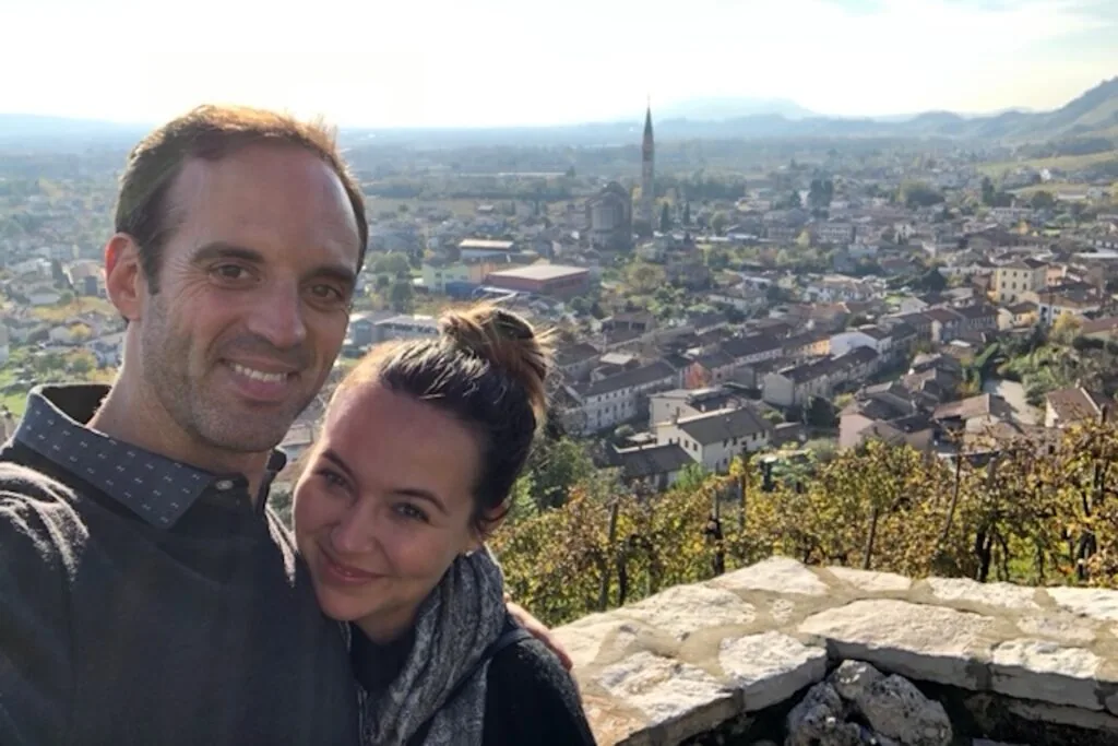 Married couple Lindsay Verber and Gregory Smith smile in selfie-style image overlooking European city.