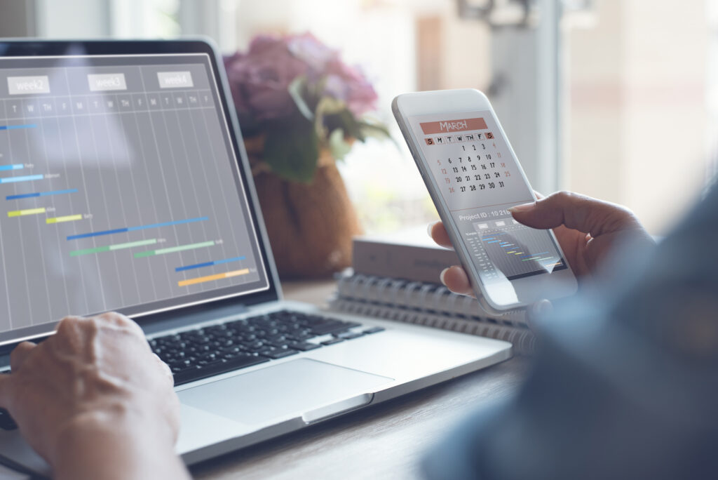 An open laptop shows a timeline while a person checks their phone calendar in the foreground. This article explains the difference between a product manager and a project manager.