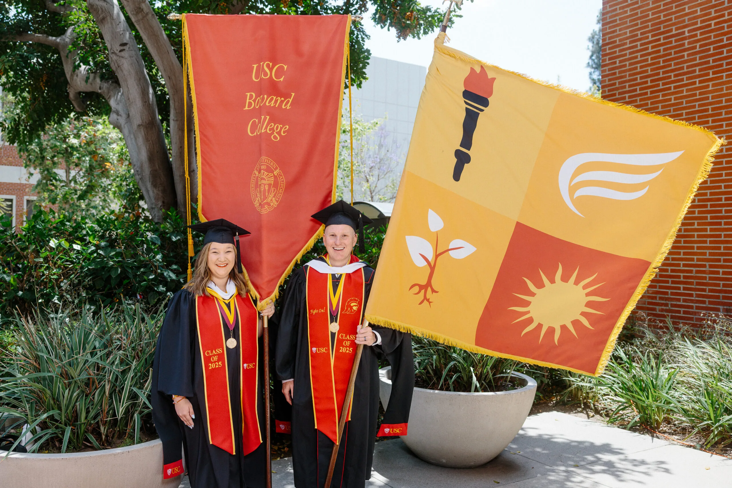 Read about: Jonathan Francisco wears graduation garments and holds a yellow flag, standing beside USC Bovard College banner bearer Devyn Favela.
